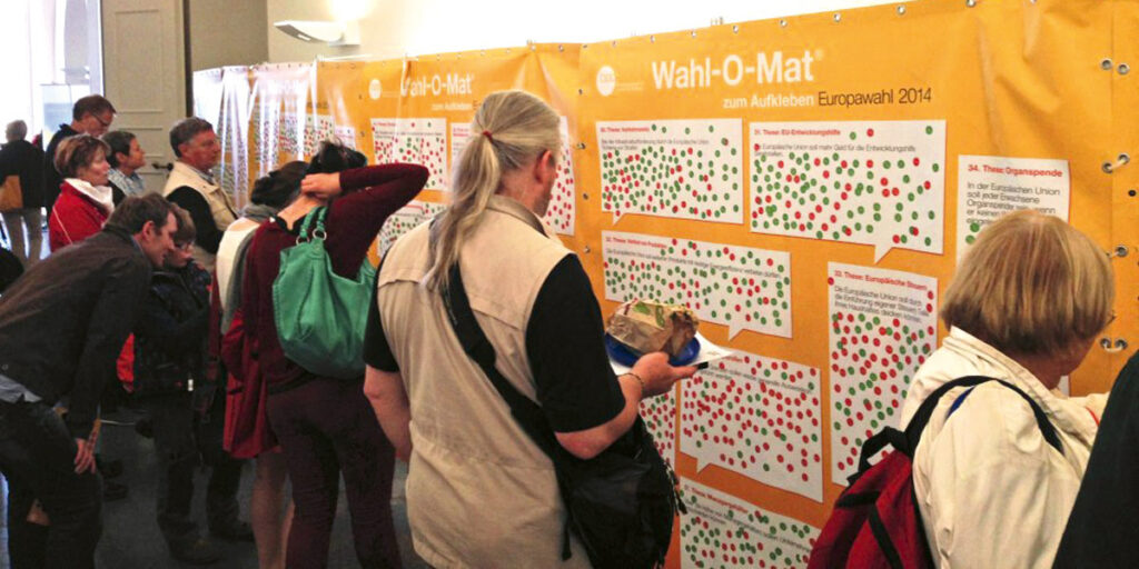 Menschen stehen vor dem Wahl-O-Mat als orangefarbene Wand mit Stickern zum Aufkleben